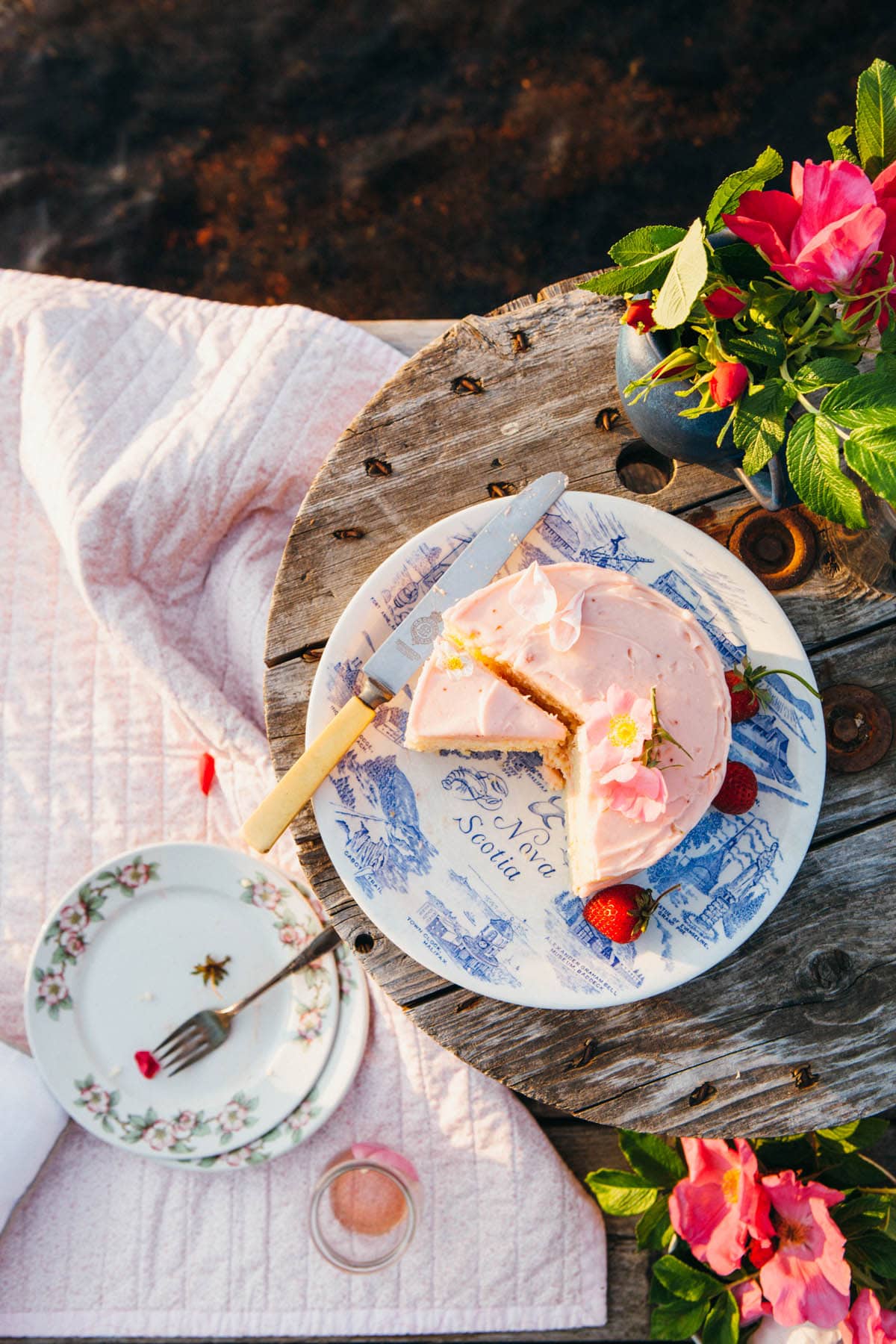 A sliced cake with pink frosting on a blue and white plate sitting on a weathered grey table on a lakeside dock.