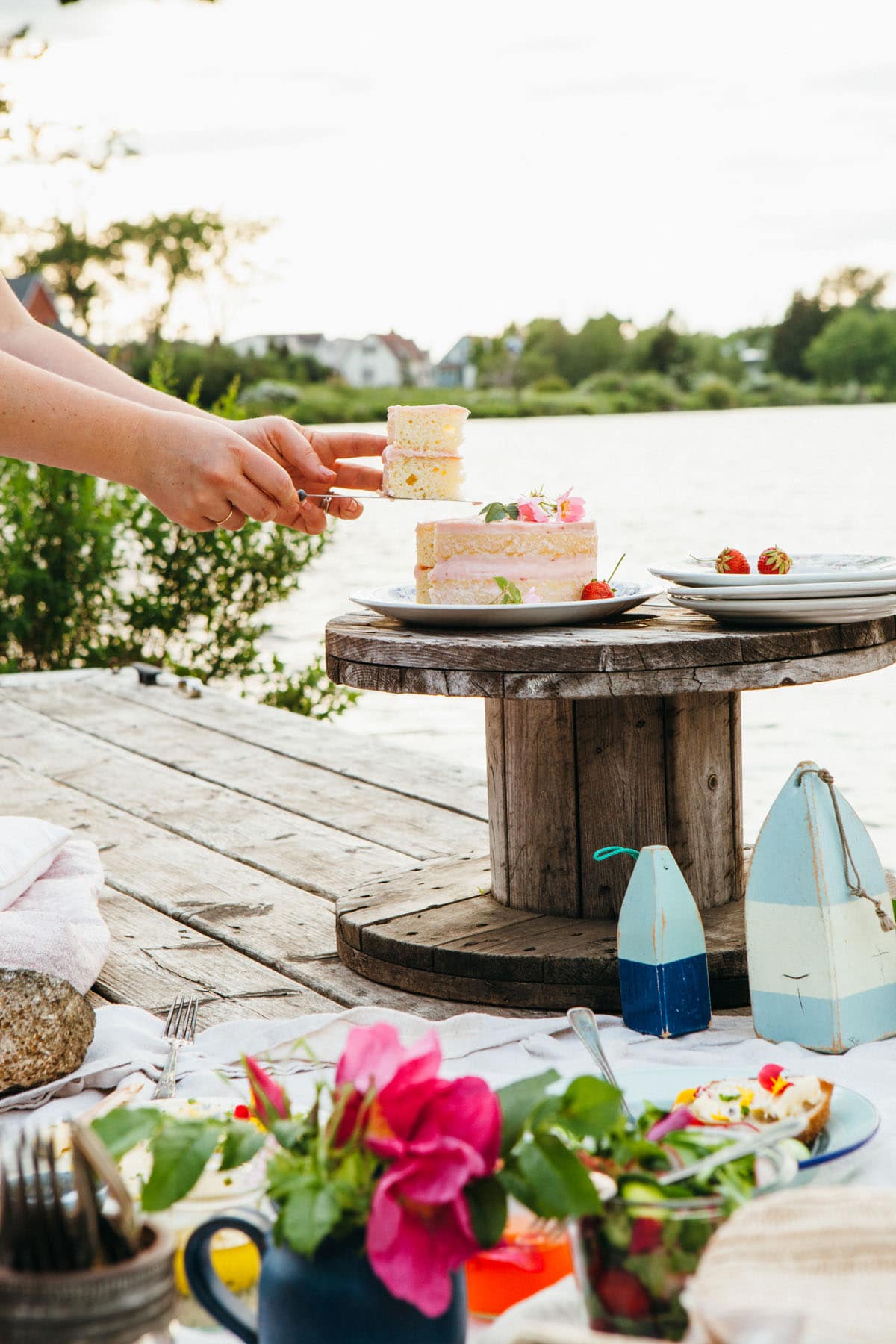 A woman's hands holding a piece of cake up after slicing it from a whole cake on a small table on a dock.