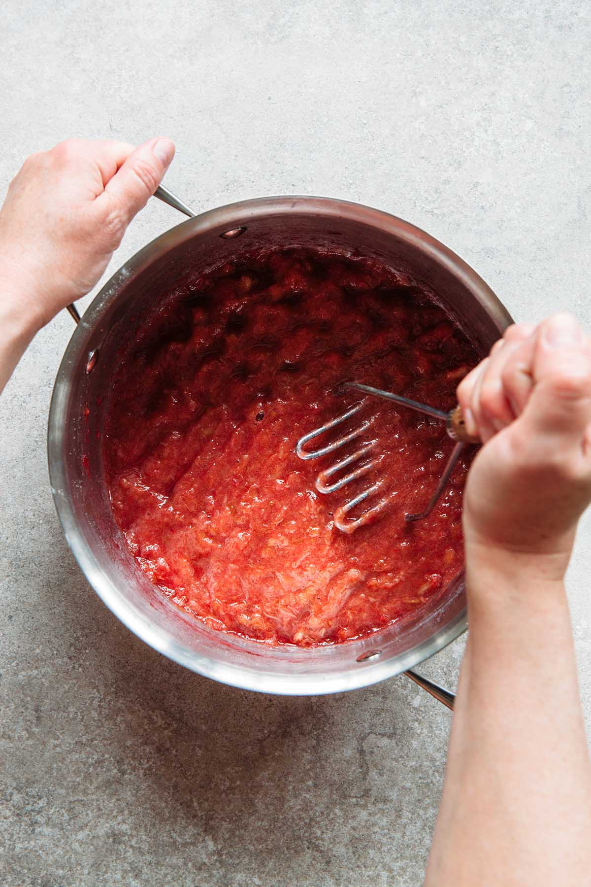 A hand crushing cooked rhubarb with a potato masher.