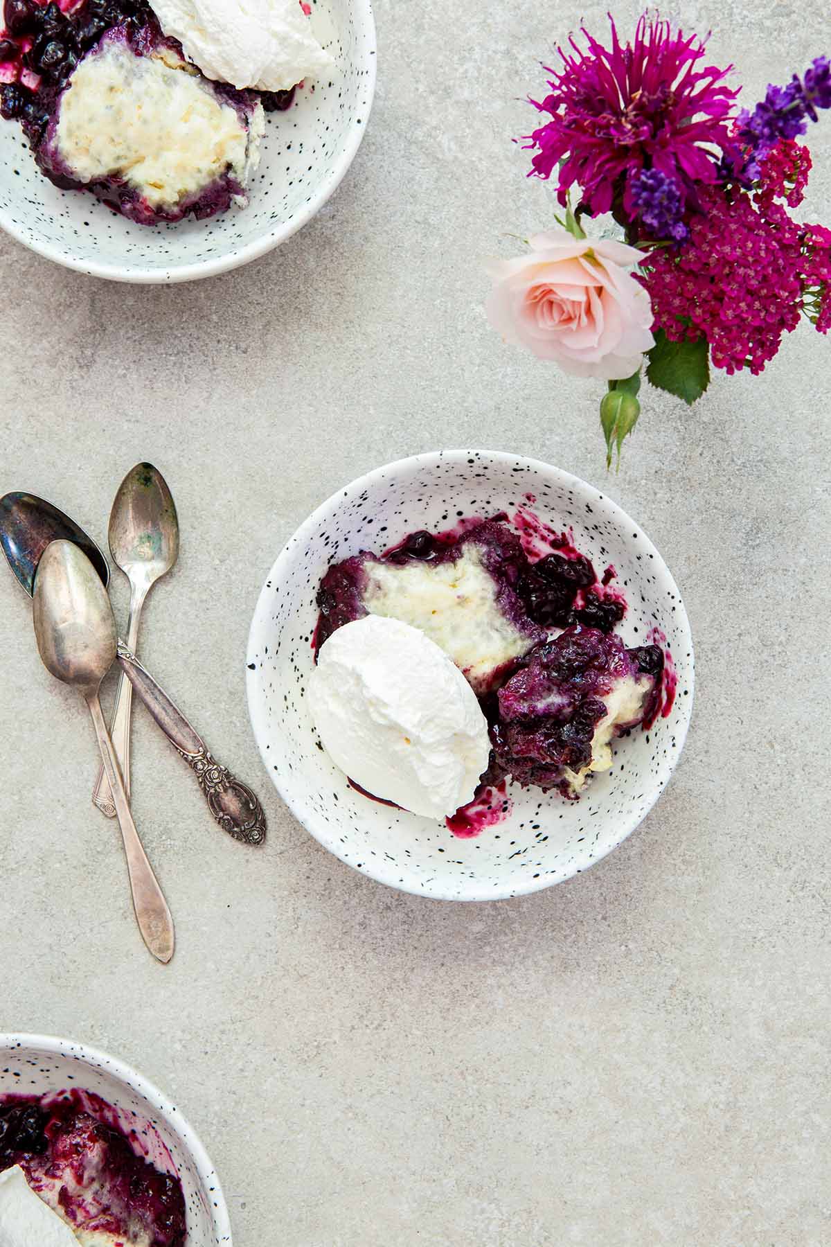Three servings of blueberry grunt with whipped cream in small speckled bowls on a stone surface next to a vase of pink and purple flowers.