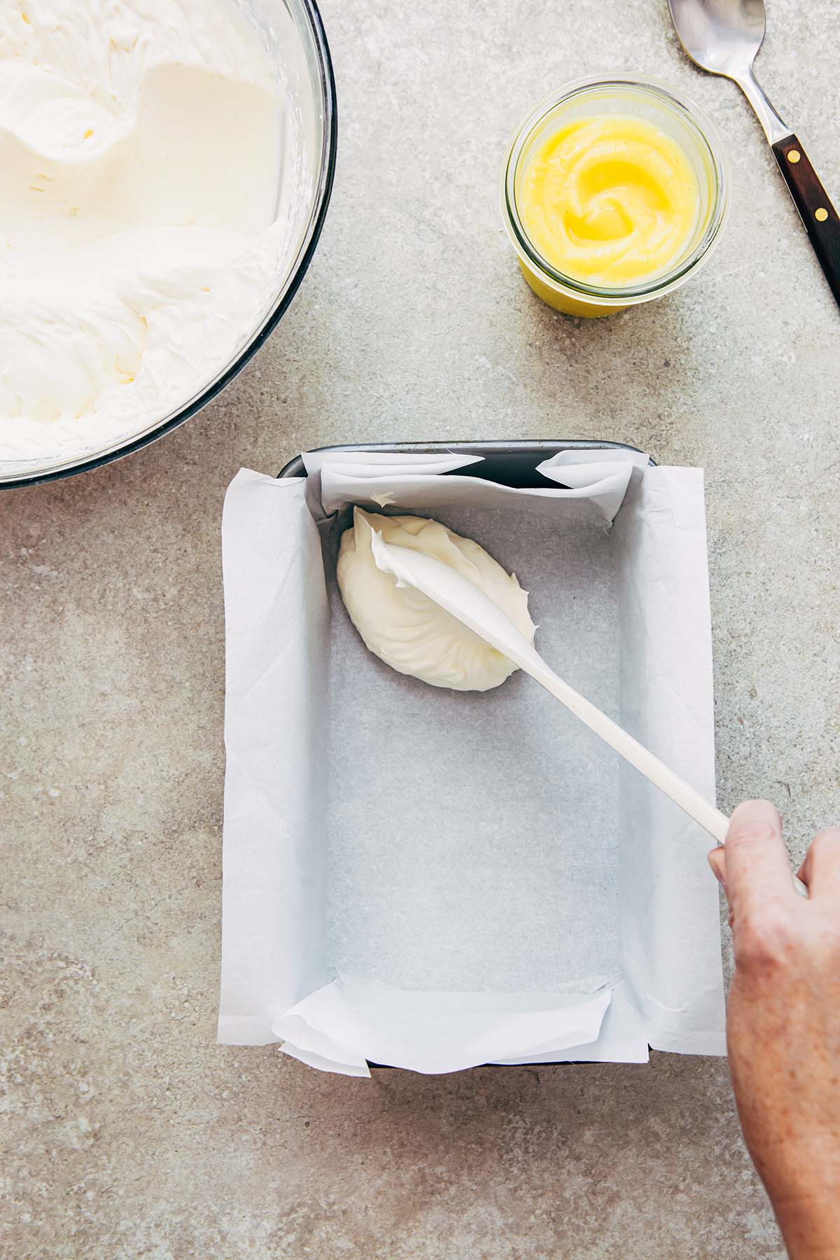 A hand dolloping whipped cream into a loaf tin lined with parchment paper.