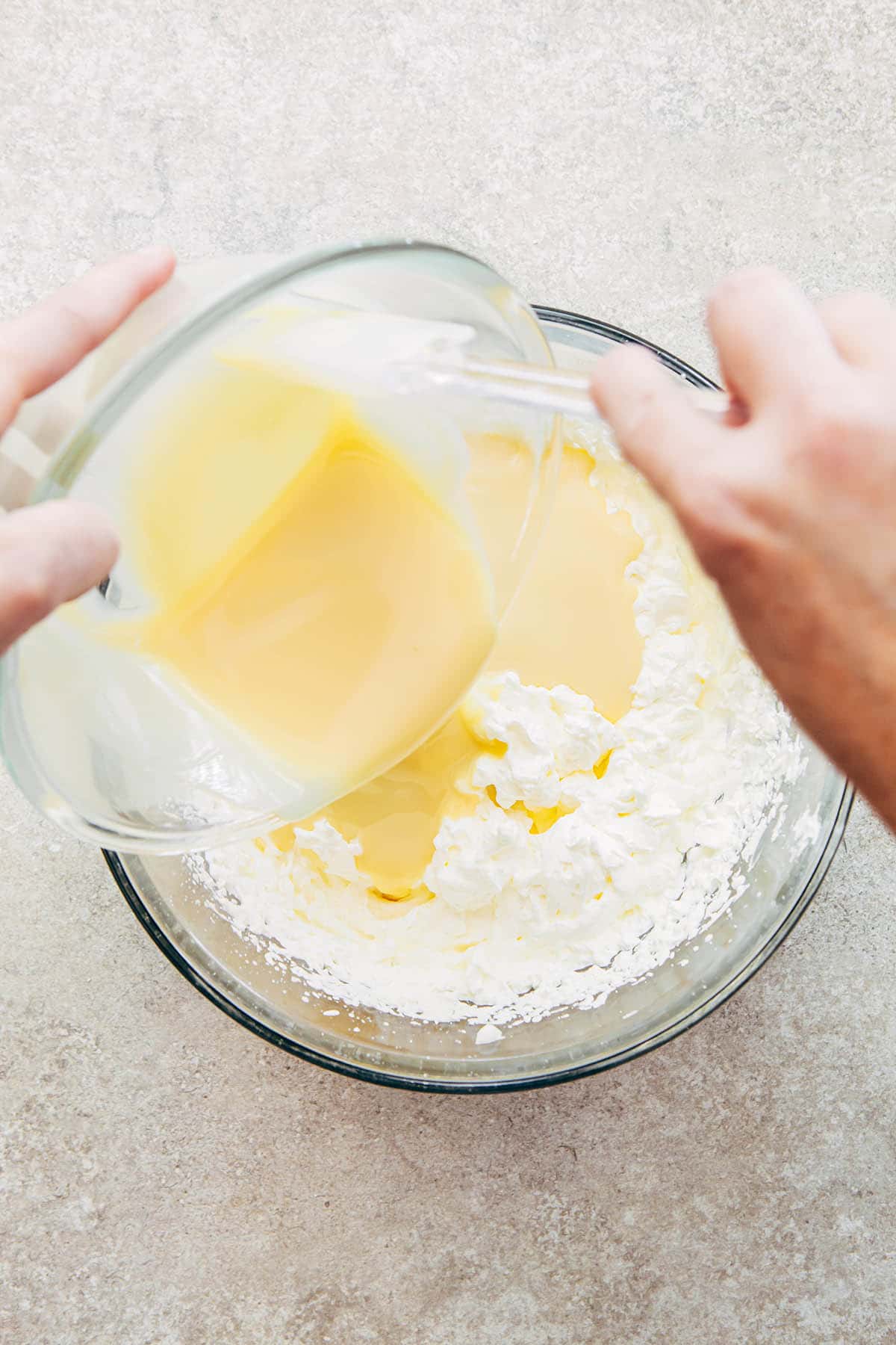 A hand using a rubber spatula to scrape condensed milk from a glass bowl into a bowl of whipped cream.