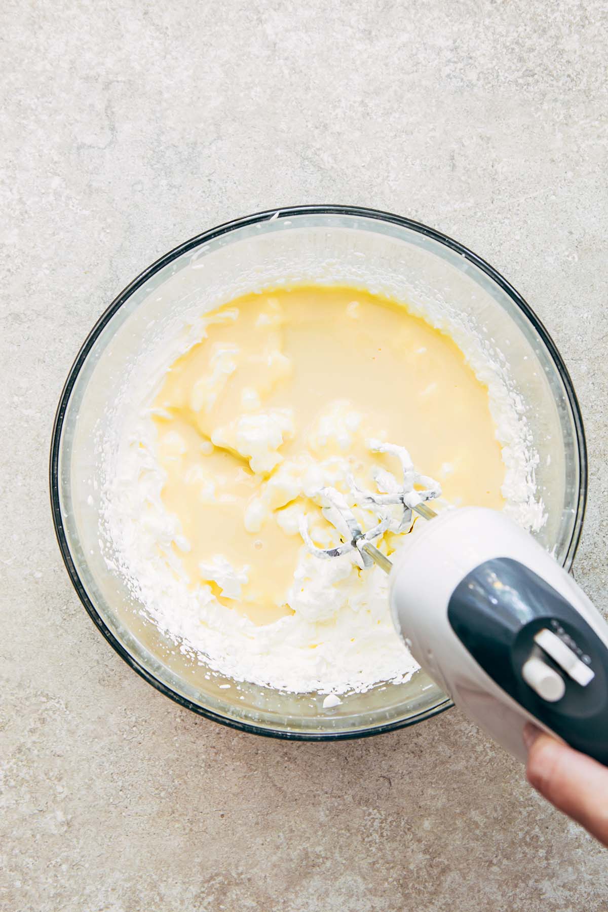 A hand mixer mixing cream and condensed milk together in a large glass bowl.