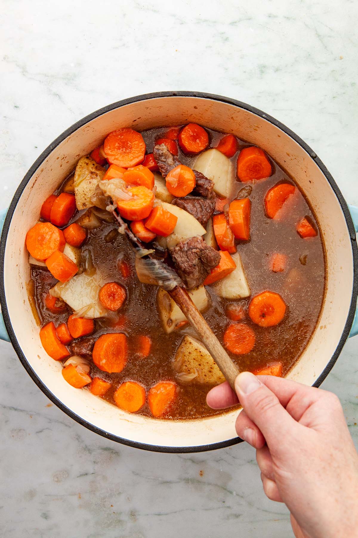 A hand using a wooden spoon to stir together cooked beef cubes, beef broth, and onions with raw potatoes and carrots.