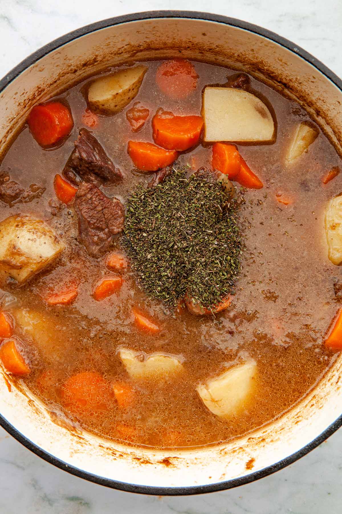 A pot of cooked beef, broth, onions, carrots, and potatoes with a small pile of dried summer savory sitting on top of the mixture.