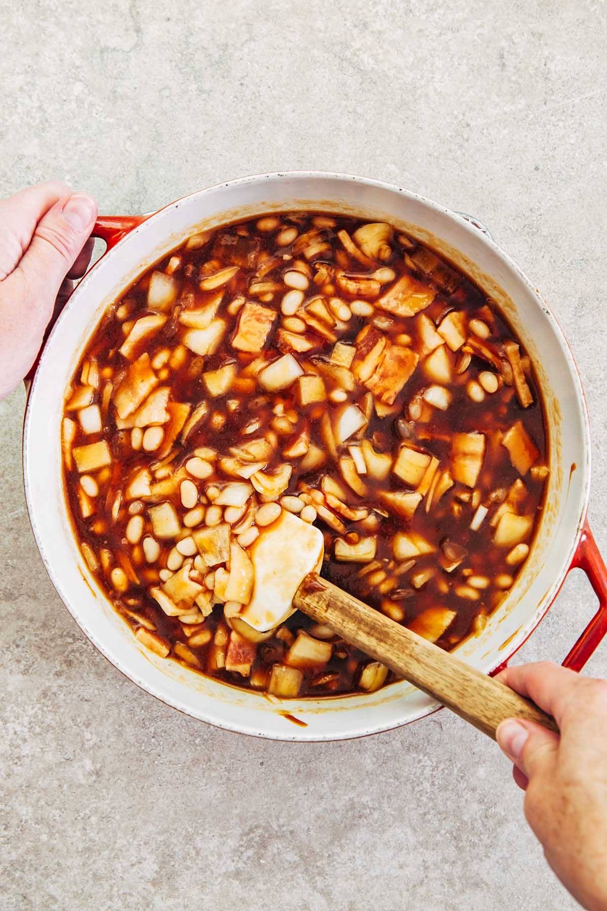 Hands stirring a pot of beans, ketchup, molasses, bacon, and onion with a wooden handled rubber spatula.