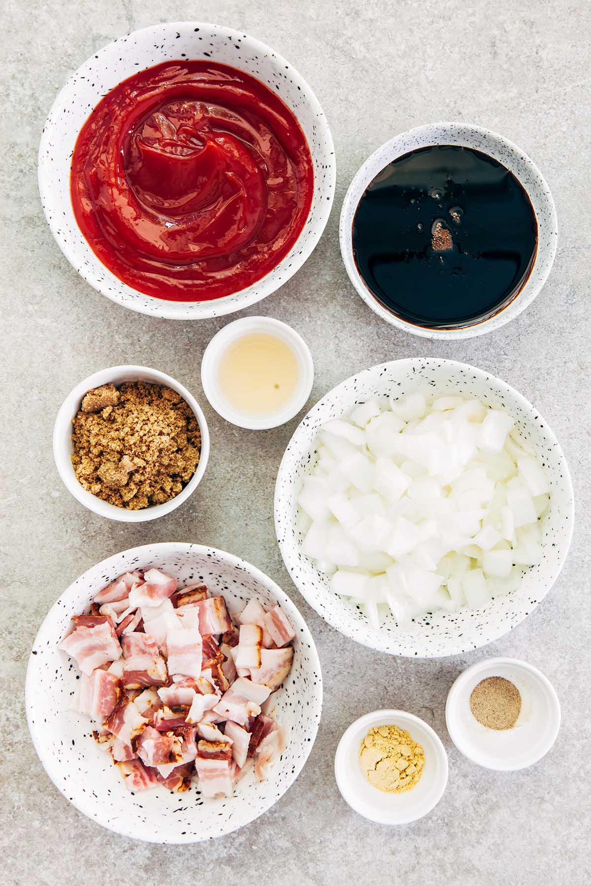 Bowls of ketchup, molasses, apple cider vinegar, brown sugar, chopped onion and bacon, ground mustard, and ground pepper on a stone surface.