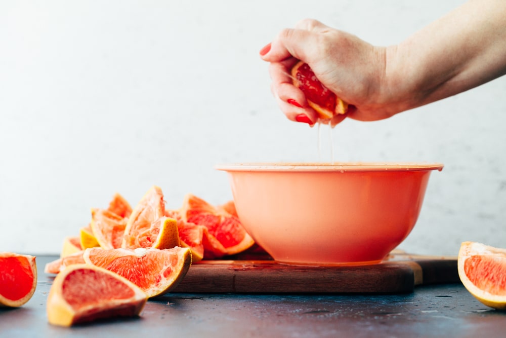 A hand over a bowl squeezing juice from a wedge of grapefruit.