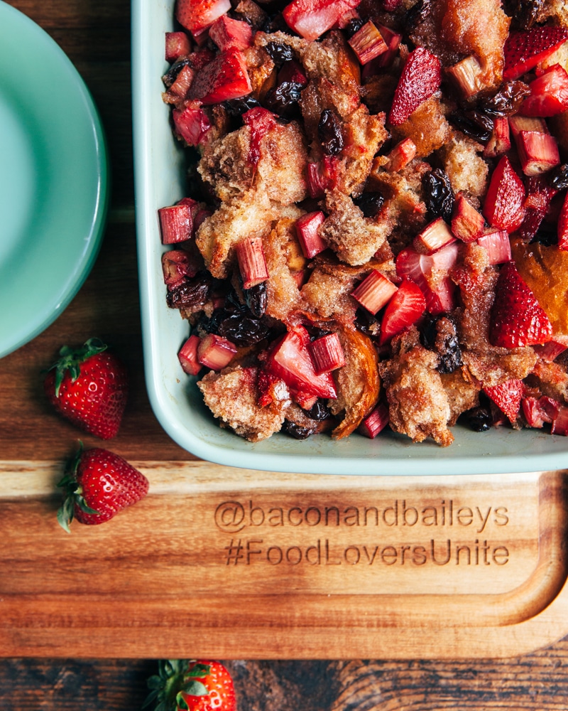 A green baking dish of strawberry rhubarb bread pudding cooling on a wooden cutting board.