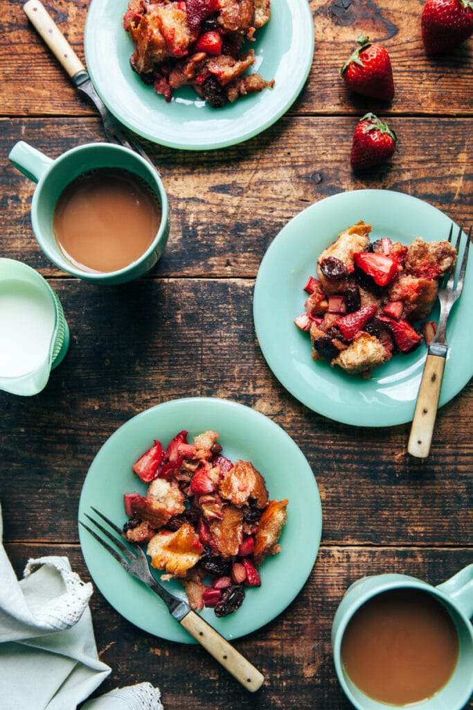 Three servings of strawberry rhubarb bread pudding on green plates with two cups of tea on a harvest table.