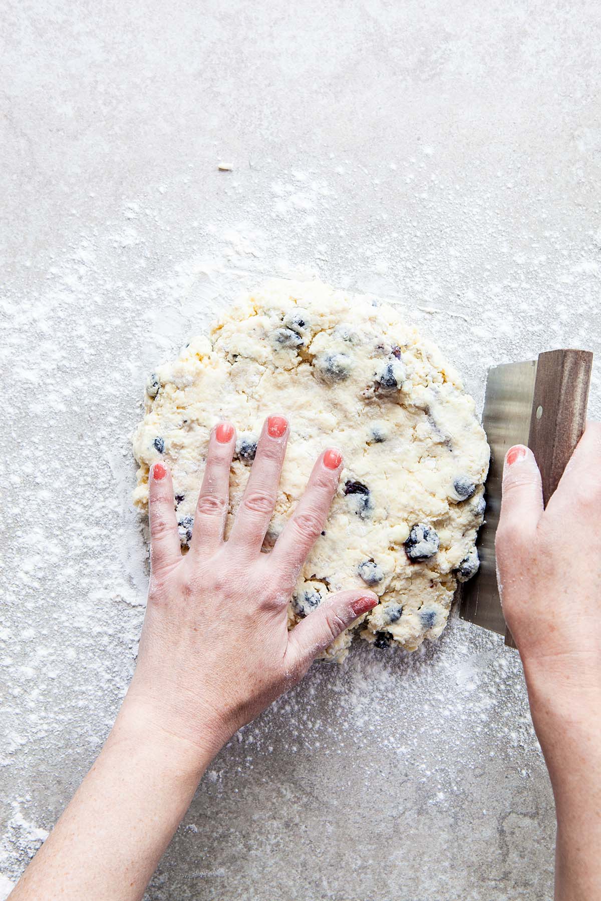 Two hands using a bench scraper to shape and smooth dough into a smooth disc on a floured stone surface.