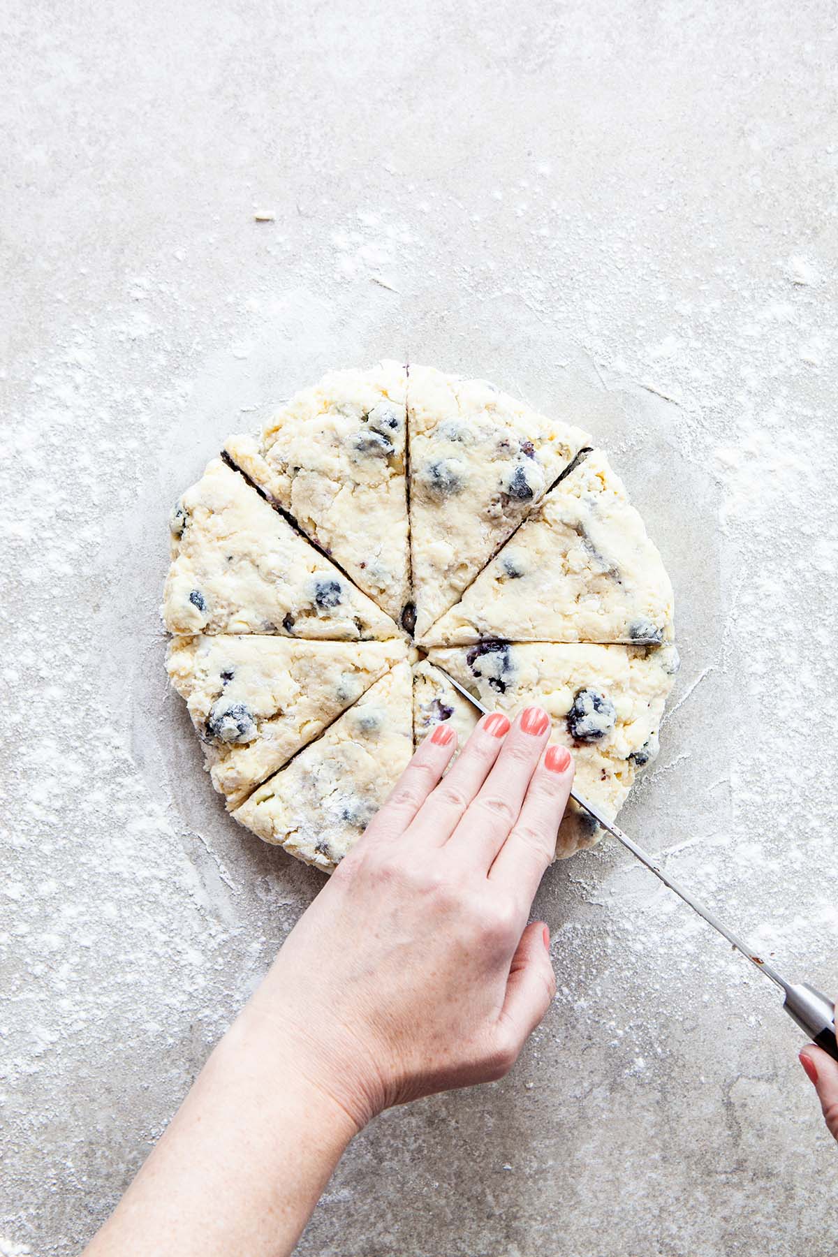 Hands cutting a disc of scone dough into wedges with a long knife on a floured stone surface.