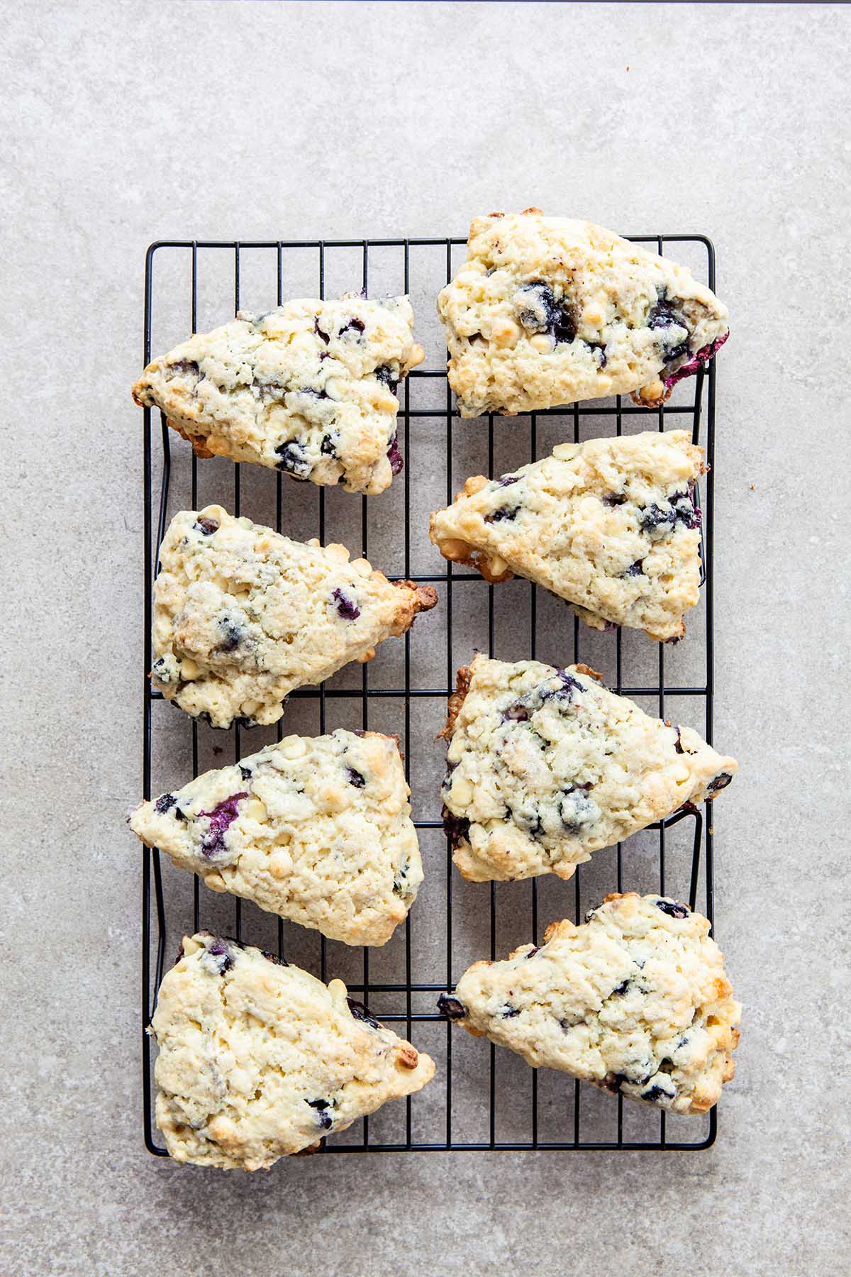 Eight blueberry white chocolate scones cooling on a rack set on a stone surface.
