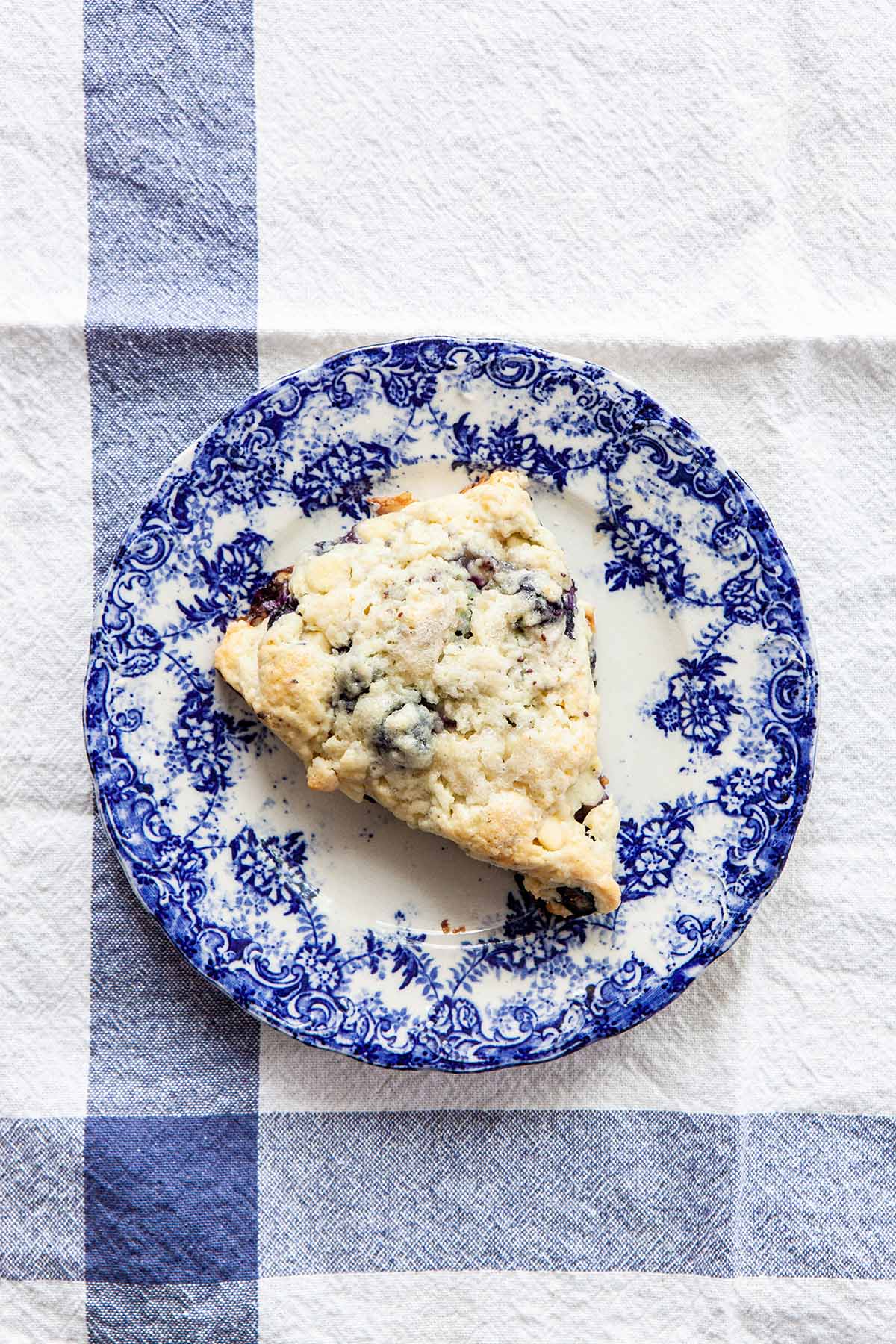 One blueberry white chocolate scone sitting on a bright blue and white plate on a white tablecloth with two blue stripes.