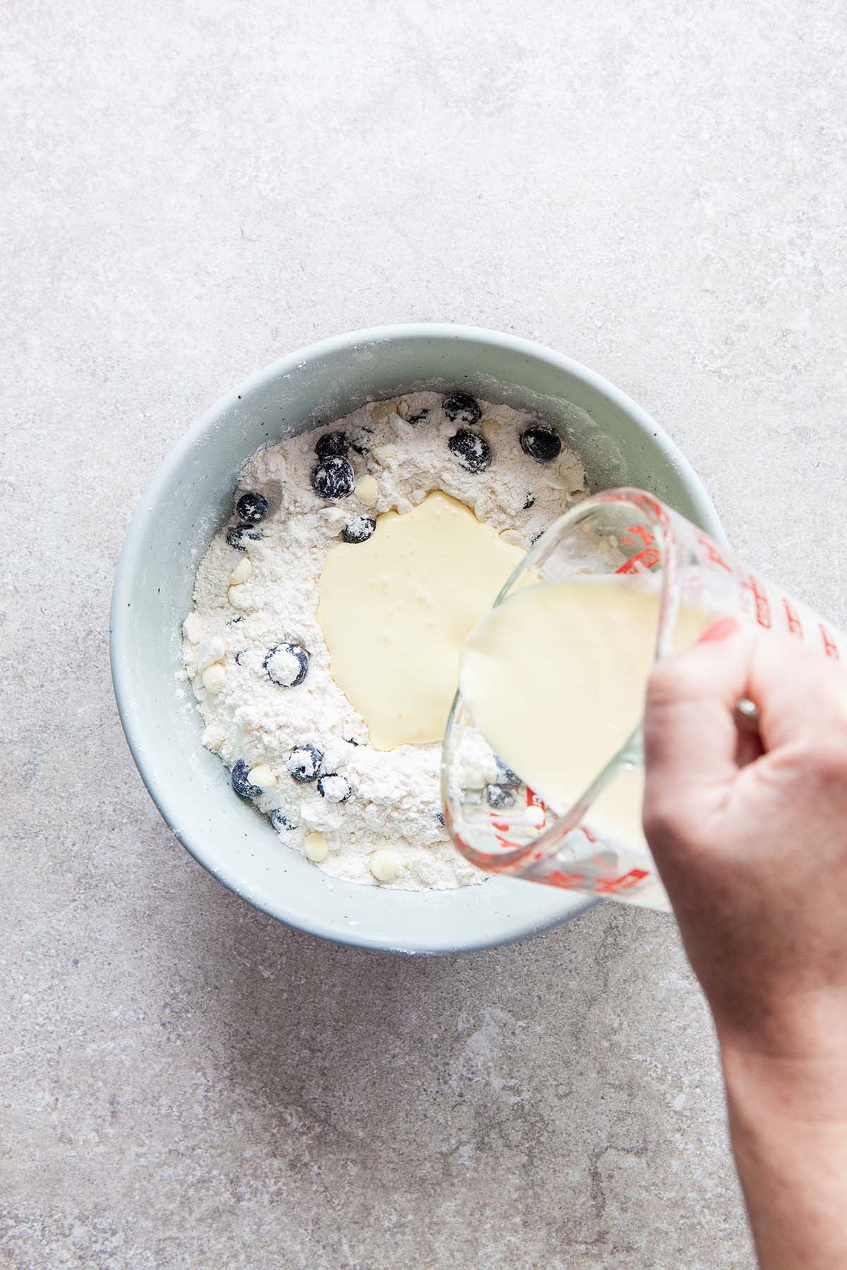 A hand pouring cream into a bowl filled with flour, blueberries, and white chocolate chips.