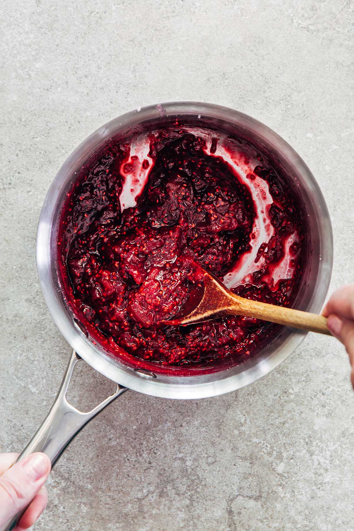 Cooked jam in a pot on a stone surface being stirred with a wooden spoon.