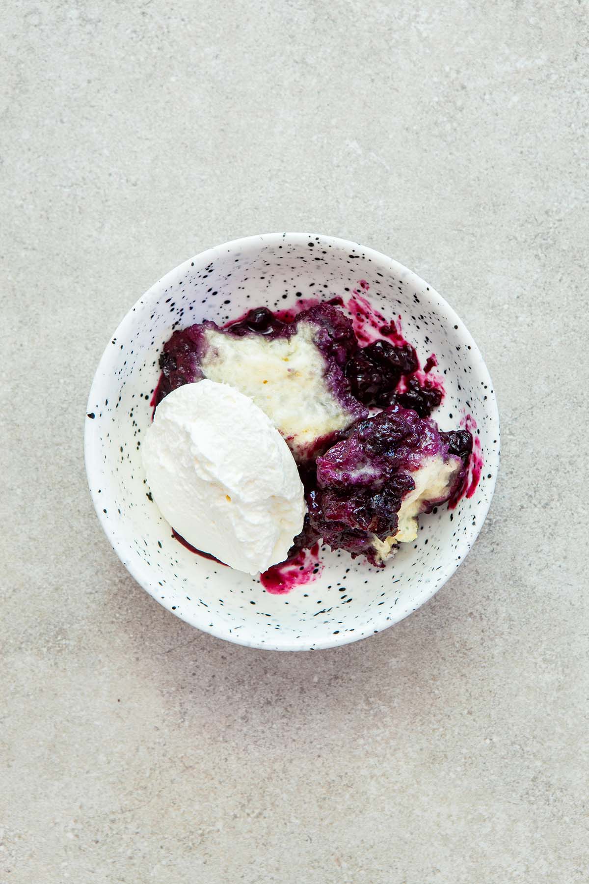 A bowl of blueberry grunt topped with whipped cream in a small white speckled bowl.