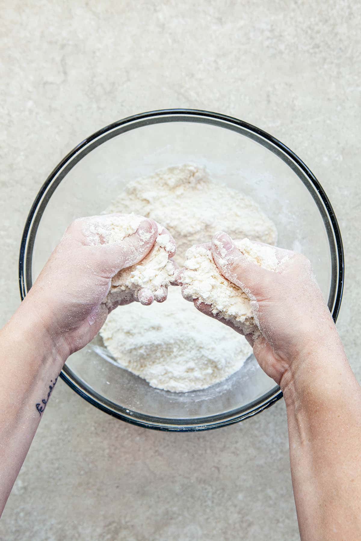 Two hands rubbing butter into flour in a glass bowl.