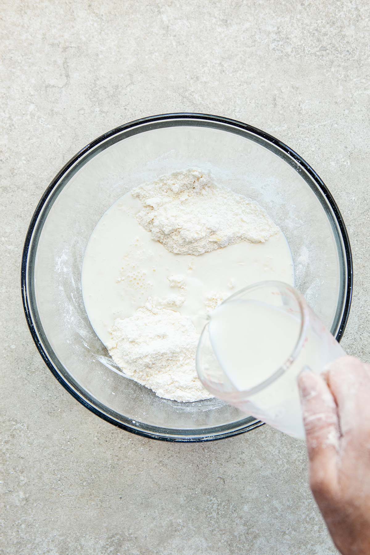 A hand pouring milk into a bowl of dry biscuit ingredients.