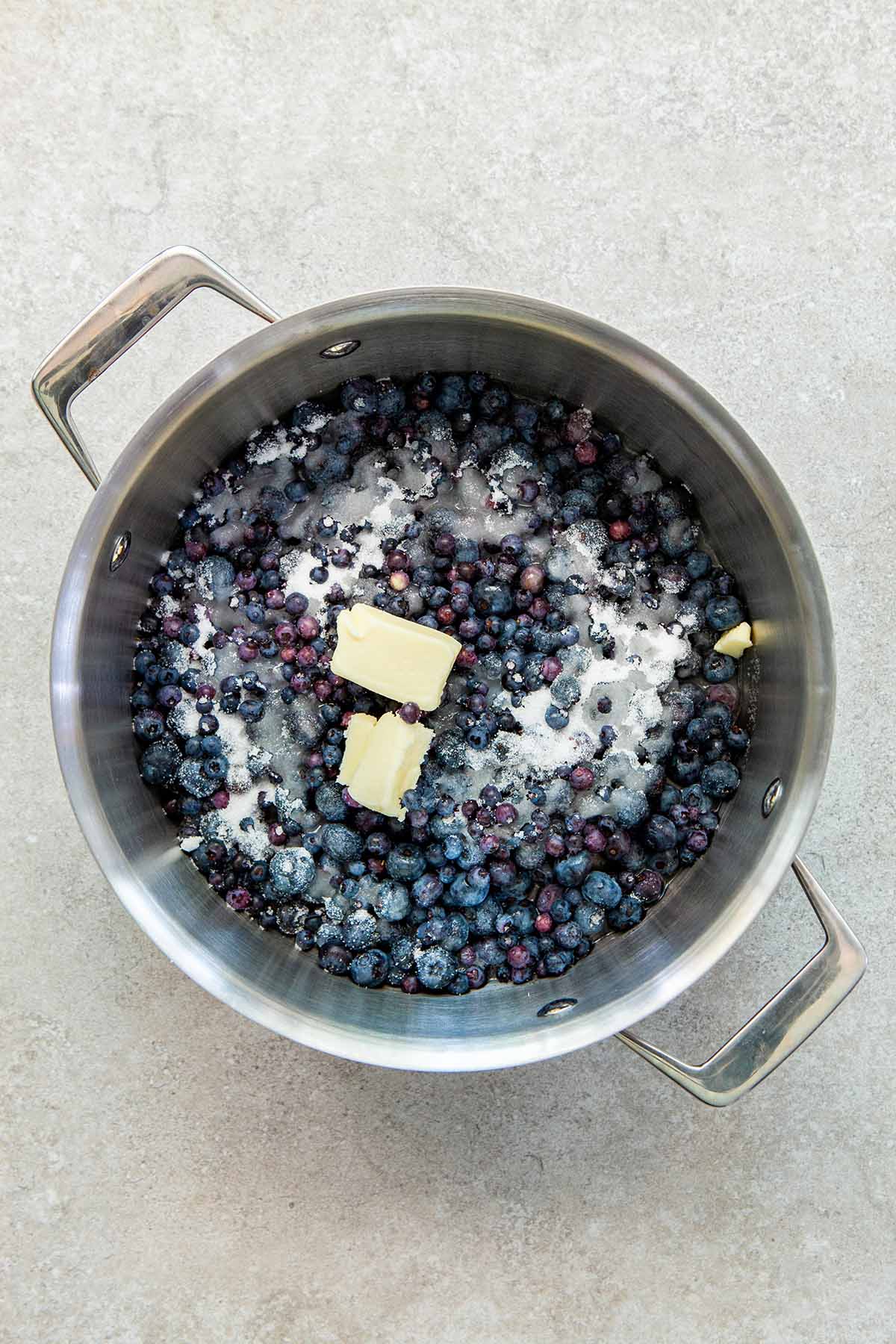 A silver pot with handles with wild blueberries, sugar, water, and chunks of butter inside.