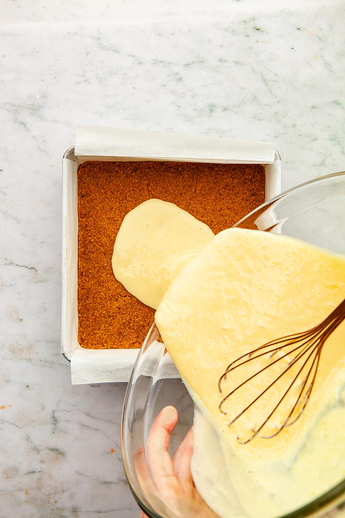 Hands pouring lemon filling from a large glass bowl into a square pan lined with graham crust.