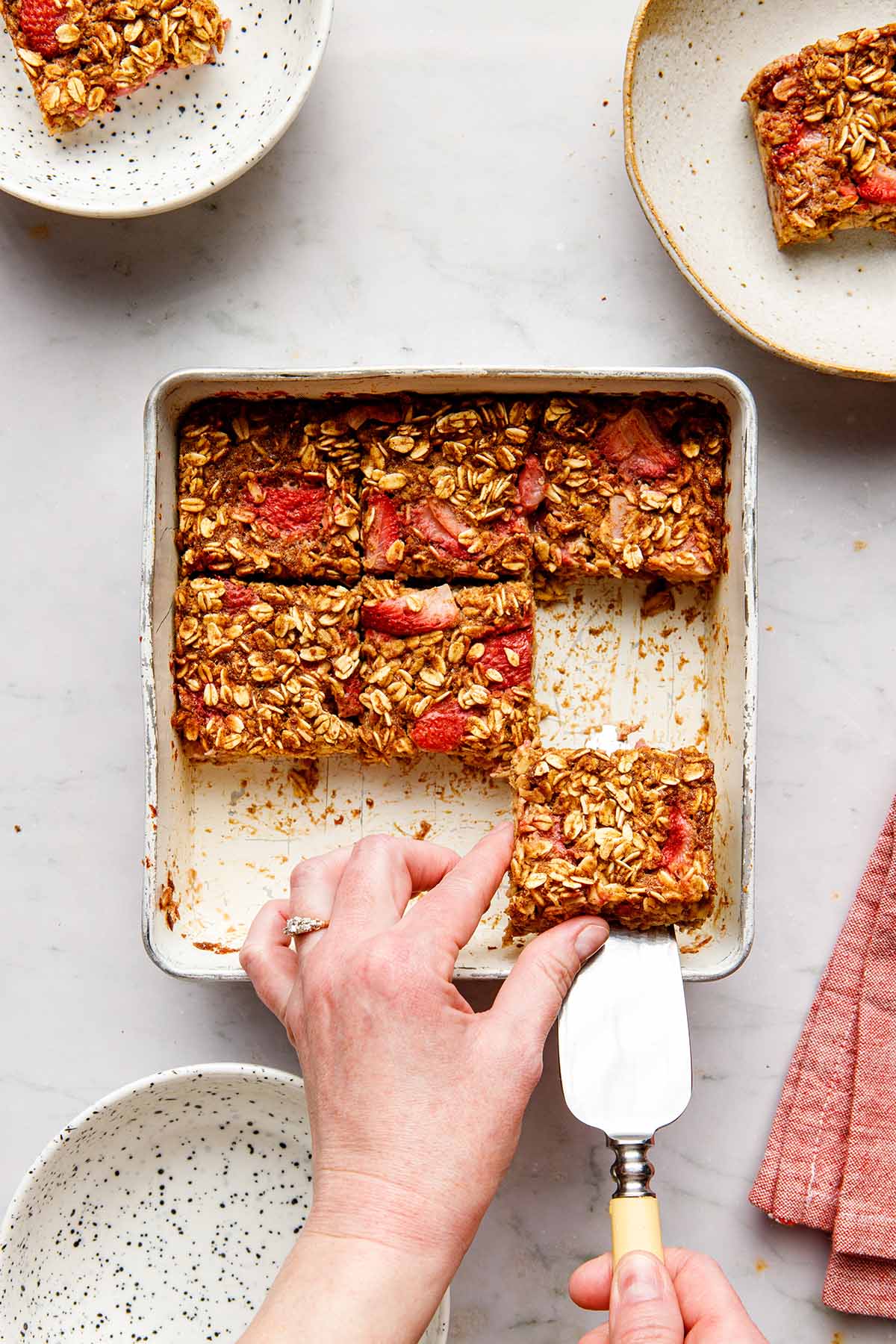 A hand using a pie lifter to lift a square slice of vegan strawberry baked oatmeal from a square baking tin.