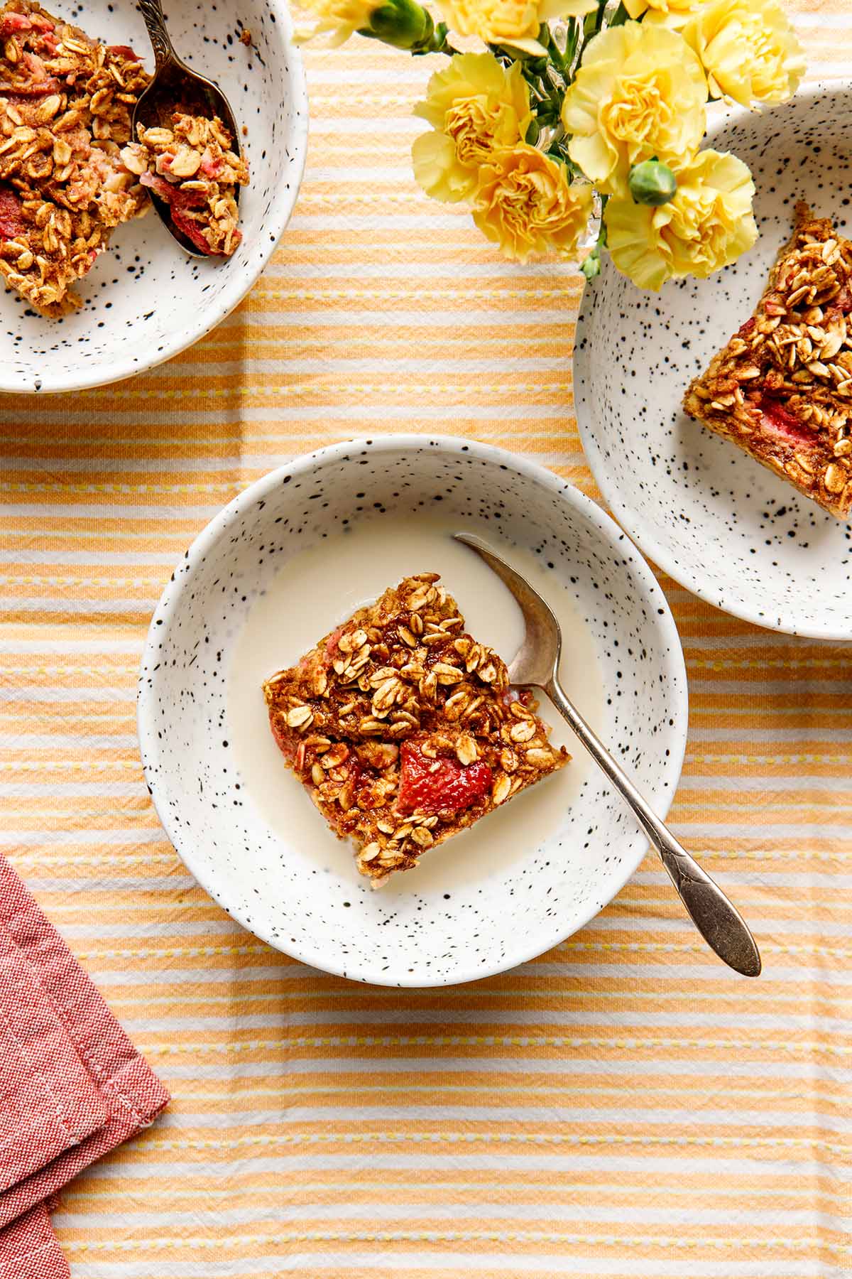 Servings of vegan strawberry baked oatmeal, topped with plant-based milk and maple syrup, in bowls on a table with a striped orange tablecloth.