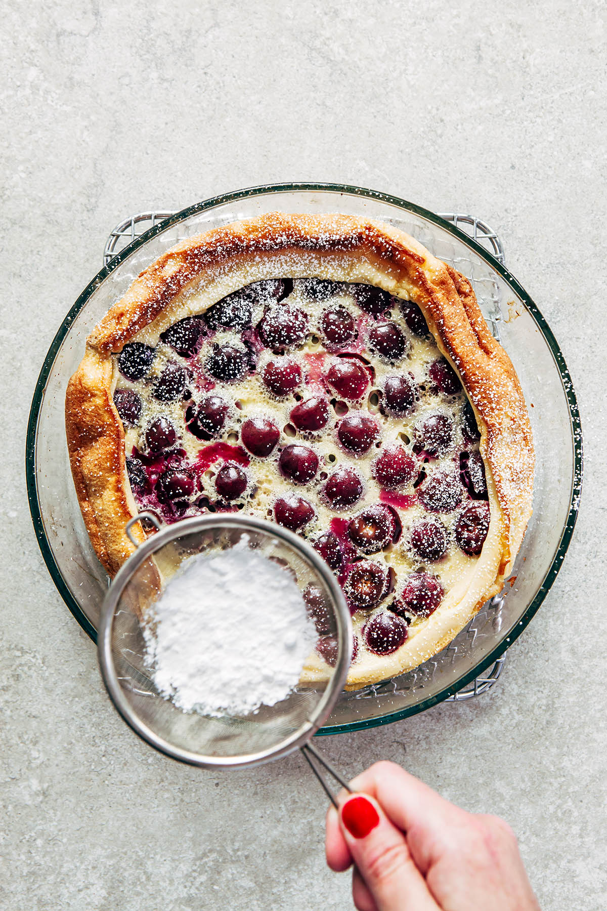A hand sprinkling powdered sugar through a small sieve over top of a baked cherry clafoutis in a round glass baking dish.