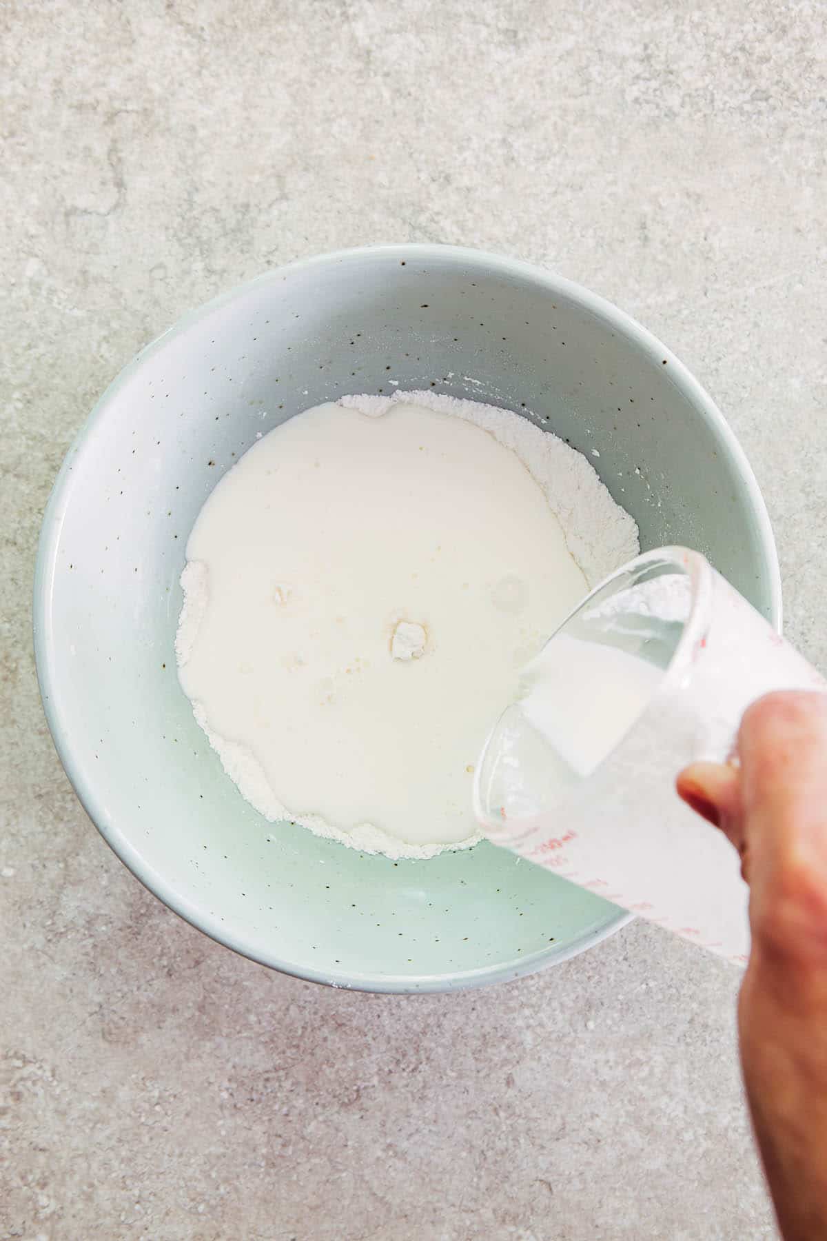A hand pouring milk from a measuring cup into a bowl of flour.