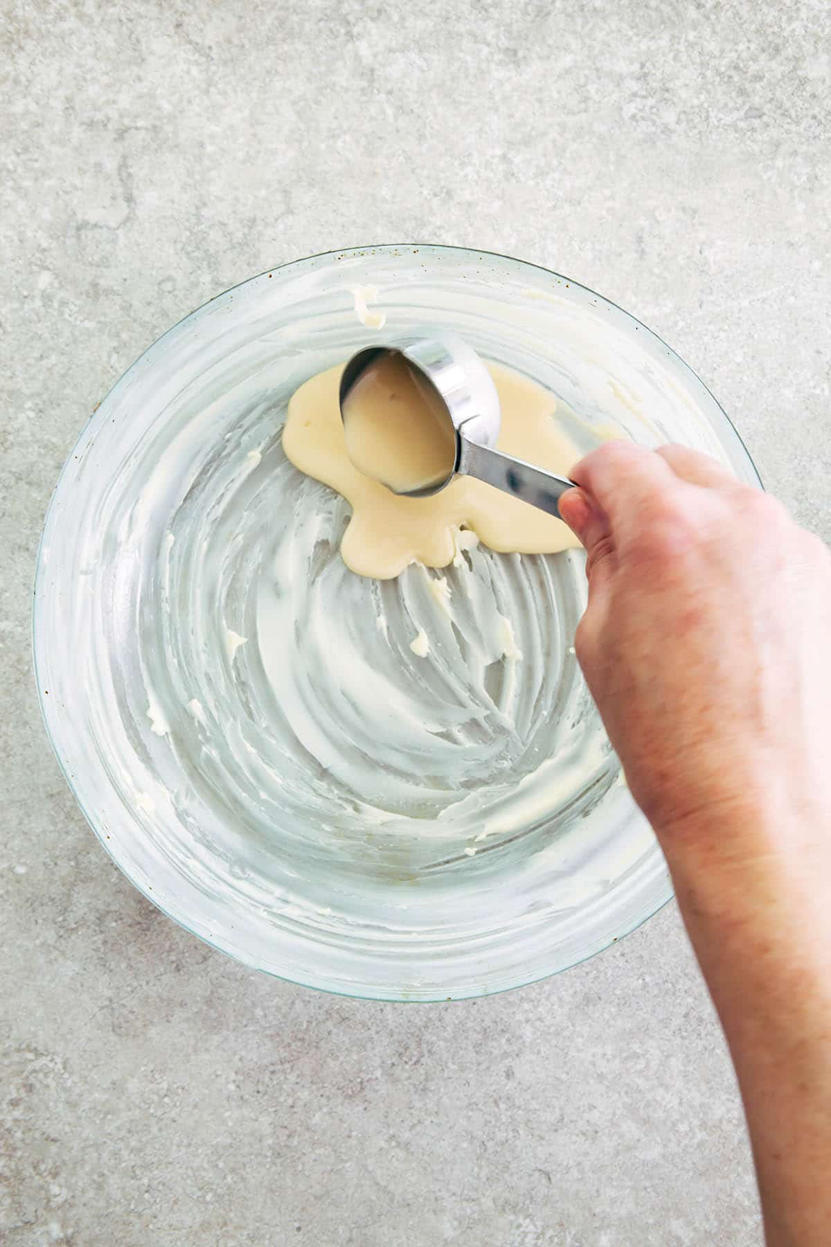 A hand pouring a small amount of batter from a small measuring cup into a round buttered baking dish.