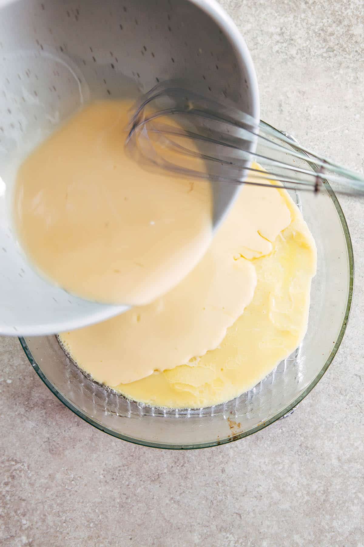 A bowl of batter being held over a baking dish with the batter being coaxed out of the bowl with a small whisk.