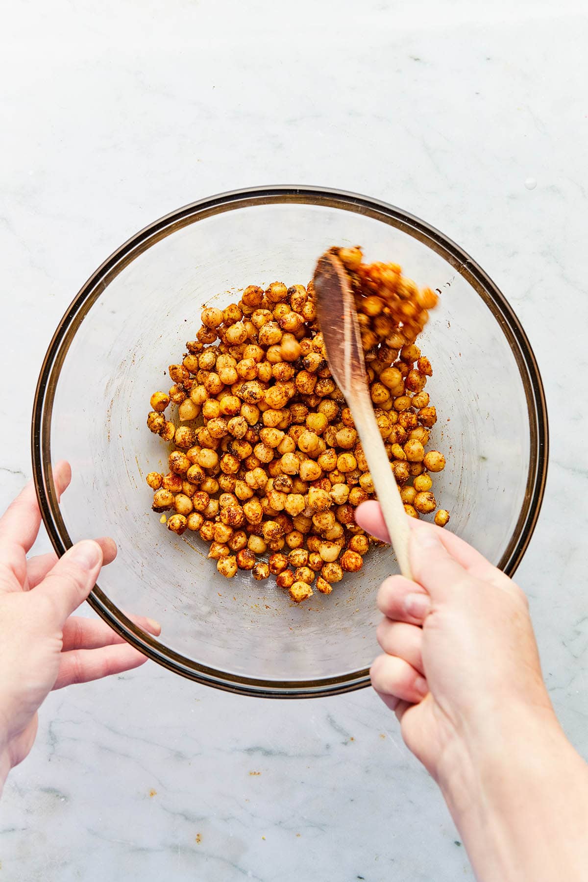 A hand stirring chickpeas and spices in a glass bowl using a wooden spoon.