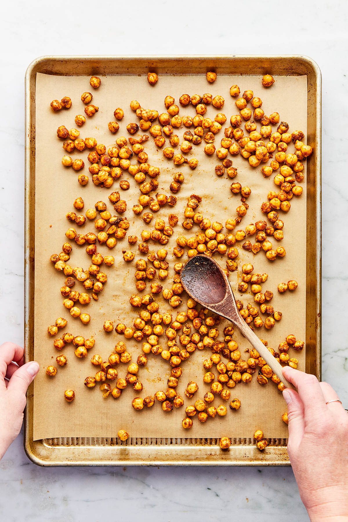 A hand using a wooden spoon to spread uncooked spiced chickpeas evenly on a baking sheet for roasting.