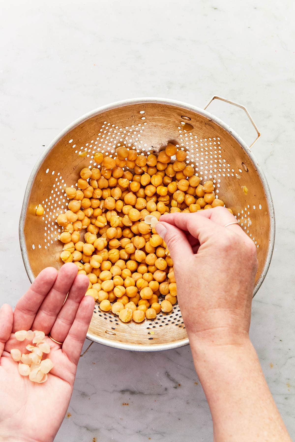 Hands plucking loose shells from rinsed chickpeas in a colander.